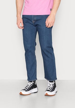 Mand iført blå straight-leg jeans, sort-hvide høje sneakers og en pink skjorte, der er kortet over taljen.