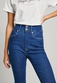 Högmidjade blå jeans med en slät denimtextur, som har två framfickor, silverknappar och gula sömnadsdetaljer.