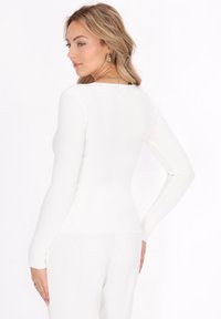 Maglietta bianca a maniche lunghe a costine con un design aderente. Dettagli notevoli includono un collo rotondo e una texture liscia, mostrata da un angolo laterale.