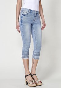 Jeans bleu clair coupés avec ourlets brodés et bords effilochés, portés avec des sandales à talons hauts noires et beiges sur une personne debout.