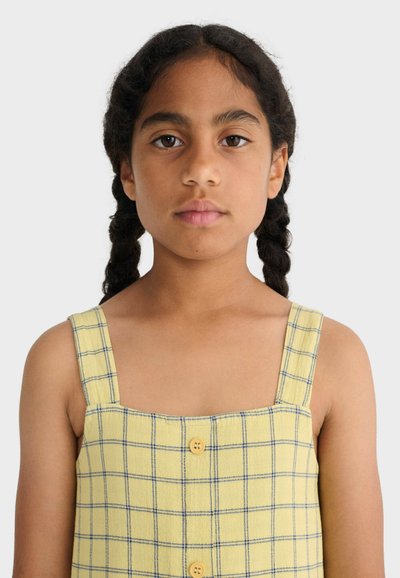 Jeune fille aux cheveux tressés portant une robe jaune sans manches à motif à carreaux bleus avec des boutons, vue de face sur un fond uni.