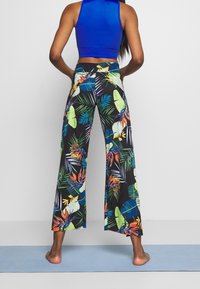 Blommiga vida byxor i svart med livfulla gröna och orange tropiska bladmönster; kombinerat med en enfärgad blå ärmlös topp.