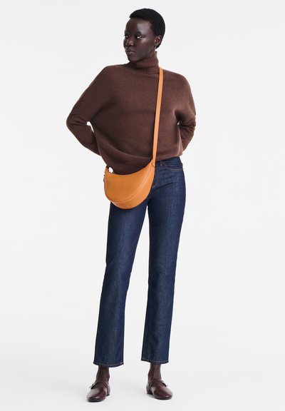 Brauner gestrickter Rollkragenpullover, dunkelblaue gerade Jeans und eine beigefarbene Umhängetasche. Model trägt braune Loafers. Schlichter Hintergrund.