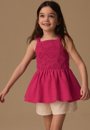 Niña con cabello largo y oscuro que lleva un vestido sin mangas de color rosa brillante con bordados florales y pantalones cortos beige sobre un fondo beige liso.