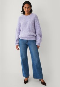 Vrouw in een lichtpaarse fluffy trui, blauwe wijde jeans, zwarte puntige hakken en zilveren ringoorbellen tegen een effen achtergrond.