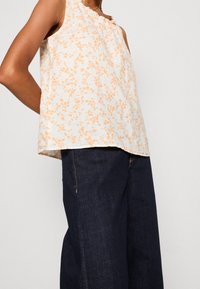 Blouse légère avec un col à volants, motif floral orange sur fond blanc, portée avec un jean large bleu foncé. Tissu doux et lisse.