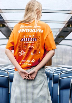 Persoon met lang blond haar staat in een bus of trein en draagt een oranje shirt met een grafiek van een bloemenwinkel in Amsterdam en een geruite rok.