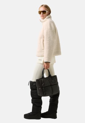 Frau in einer cremefarbenen Fleecejacke, weißen Leggings, schwarzen gepolsterten Stiefeln, großen Sonnenbrillen und einer schwarzen gepolsterten Tragetasche.