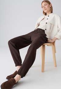 Vrouw zittend op een houten stoel, gekleed in een crèmekleurig jasje met knopen, een wit shirt, donkerbruine joggingsbroek en bruine instapschoenen.