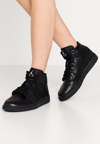 Mustat korkeat tennarit, joissa on teksturoitu nahka, näyttävät nauhat, kumipohjat ja logo kielekkeellä. Suunniteltu tyyliä ja mukavuutta varten.