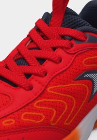 Κοντινό πλάνο κόκκινου αθλητικού παπουτσιού με υφασμάτινη υφή, κόκκινες κορδόνια με σχέδια, πορτοκαλί λεπτομέρειες και μαύρο-γκρι λογότυπο στο πλάι.