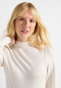 Cremefarvet sweater med høj ribkrave, lange ærmer og et blødt, tekstureret stof. Har let samlede skuldresømme.