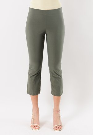 Donna che indossa pantaloni corti verde oliva e sandali beige con tacco alto, in piedi contro uno sfondo bianco uniforme.