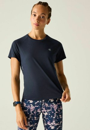 Young woman with braided hair wears a dark blue short-sleeve shirt and blue floral patterned leggings, holding the shirt hem on a plain background.