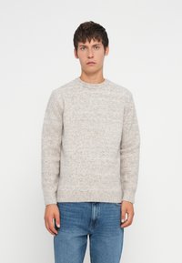 Jeune homme debout, portant un pull en tricot gris clair et un jean bleu, devant un fond blanc uni.