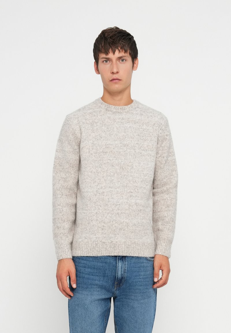 Jeune homme debout, portant un pull en tricot gris clair et un jean bleu, devant un fond blanc uni.