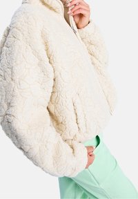 Kremowa, puszysta kurtka z zamkiem z przodu, wysokim kołnierzem i teksturowanym wzorem. Posiada boczne kieszenie i jest zestawiona z miętowymi spodniami.