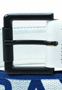 Fibbia in metallo nero su un cinturino bianco testurizzato con accenti blu. Il cinturino presenta un motivo intrecciato, che ne esalta la texture visiva.