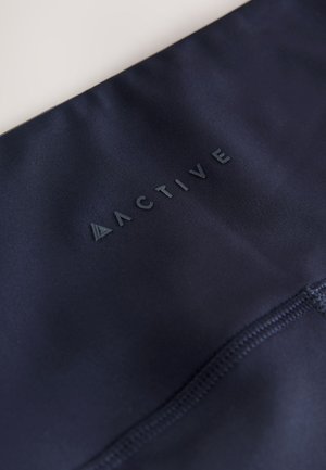 Temno modra športna oblačila z gladko teksturo, na katerih je dvignjen logotip "ACTIVE" v kontrastni temni barvi in vidni detajli šivov.