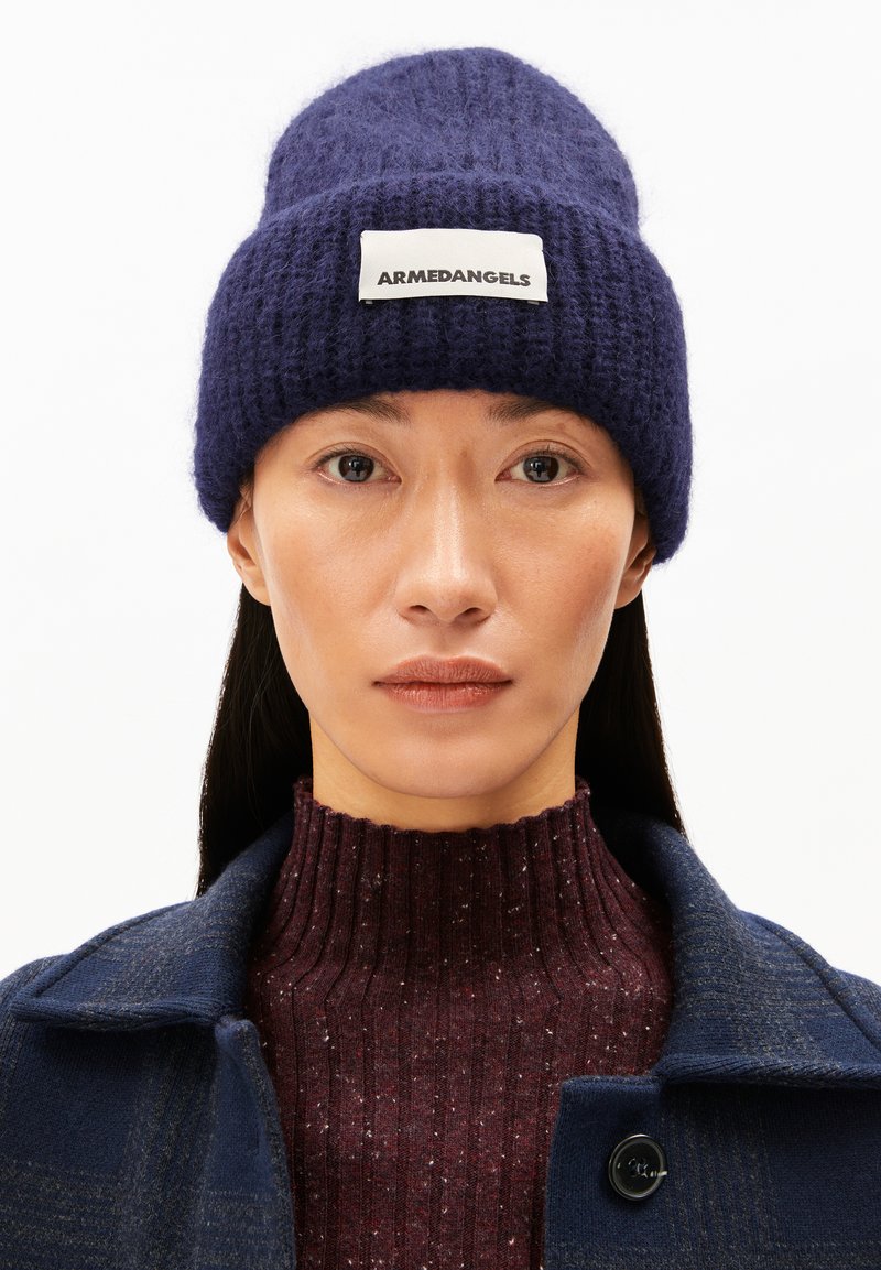 Marineblauer Strickmütze mit umgeschlagenem Bund und gesticktem Logoetikett. Kombiniert mit einem bordeauxrot melierten Pullover unter einer dunklen karierter Jacke.