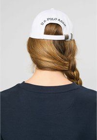 Balta beisbolo kepurė su reguliuojamu dirželiu, su juodais siuvinėjimais "U.S. POLO ASSN." Nešiojantis turi ilgus, pintus plaukus.