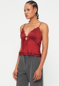 Burgundowa satynowa koszulka na ramiączkach z koronkowymi akcentami, głębokim dekoltem w kształcie litery V i regulowanymi ramiączkami. Posiada zawiązywany przód oraz warstwowy plisowany dół.