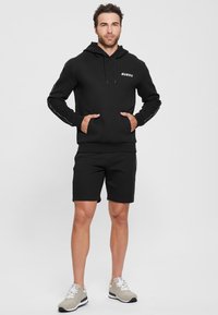 Black hoodie with front pockets and logo, paired with matching black shorts. Features grey sneakers with white accents and laces.