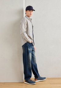 Camisa de manga larga a cuadros en azul y amarillo, pantalones vaqueros azules desgastados y zapatos navy con acentos marrones; de pie contra una pared clara.