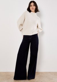 Kremowy sweter dzianinowy z teksturowanym wzorem i szerokimi rękawami, zestawiony z wysokim stanem szerokimi, ciemnoniebieskimi dżinsami.