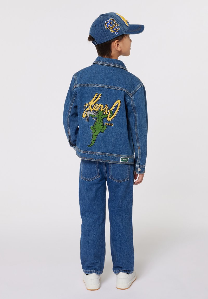 Ensemble en denim comprenant une veste et un pantalon de couleur bleu moyen. La veste arbore un grand graphique de dinosaure vert avec le texte 'Kenzo Paris' en jaune. Baskets blanches.