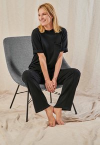 Blondes Frau in schwarzem Hemd und Hose sitzt barfuß auf einem grauen modernen Stuhl, lächelt und hält die Hände verschränkt zwischen den Knien.