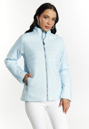 Giacca azzurra chiaro con zip frontale, colletto alto, texture trapuntata, tasche laterali e finitura in tessuto liscio. Indossata sopra pantaloni bianchi.