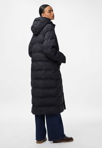 Donna che indossa un lungo piumino nero con cappuccio, jeans scuri a gamba larga e scarpe nere, in piedi davanti a uno sfondo bianco semplice.