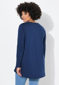 Marineblauwe lange mouw tuniek met een ronde halslijn, zachte textuur en een gebogen zoom. Gecombineerd met lichtblauwe denim, wat een losse pasvorm laat zien.