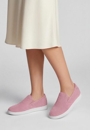 Beine, die einen cremefarbenen Rock und rosa Slip-on-Sneaker mit weißen Sohlen tragen, stehend auf einer grauen Fläche vor einfarbigem Hintergrund.