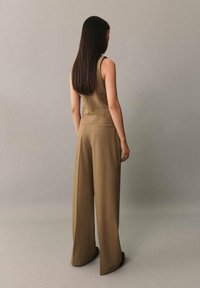 Khaki jumpsuit med en tætsiddende overdel og brede bukser. Glat stof, ærmeløs design og minimal detaljering. Model set fra bagfra.