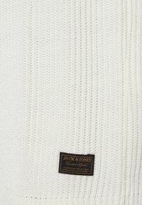 Suéter blanco de punto con textura acanalada y una etiqueta de cuero rectangular con logotipo en relieve en la esquina inferior izquierda.