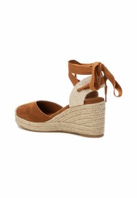 Wedge espadrille de ante marrón con suela de yute, presenta una correa de tobillo a cuadros y lazos en la parte posterior. Punta redondeada, superficie texturizada.