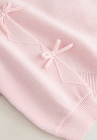 Vaaleanpunainen neulepaita, jossa on teksturoidusta kankaasta tehdyt koristeelliset rusetit ja ompeludetaljit sekä ribbineulottu hihansuu istuvampaa viimeistelyä varten.