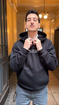 Νέος άνδρας διορθώνει το γιακά ενός μαύρου φούτερ με άσπρο κείμενο, στέκεται σε έναν θερμά φωτισμένο διάδρομο με πολυέλαιο από πάνω.