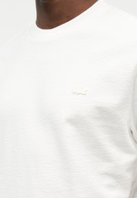 Gros plan d'une personne portant un t-shirt blanc texturé à col rond avec un petit logo de crocodile blanc sur le côté gauche de la poitrine.