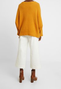 Maglione oversize senape con lavorazione a maglia texture, abbinato a pantaloni bianchi a gamba larga e stivaletti marroni con tacco a blocco.