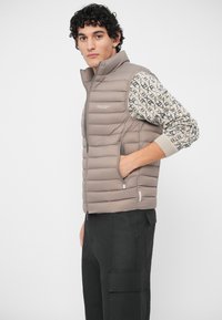 Mladić u bež prsluku s punjenjem, preko patternirane košulje dugih rukava, u crnim cargo hlačama, stoji s jednom rukom u džepu prsluka.