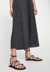 Czarne, płaskie sandały z krzyżującymi się paskami, połączone z teksturowanymi, ciemnoszarymi spodniami o szerokich nogawkach w kroku. Widoczna stopa, z otwartym przodem.