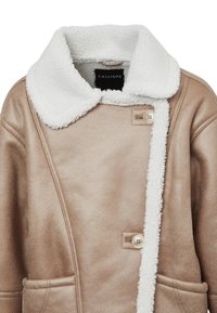 Beige-farbener Kunstpelzmantel mit einem weißen flauschigen Kragen, großen Knöpfen und einer seitlichen Tasche, der eine glatte, strukturierte Außenschicht aufweist.