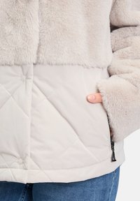 Veste légère de couleur crème avec une partie supérieure en fausse fourrure et une section inférieure matelassée. Dispose de poches latérales et d'une texture douce.