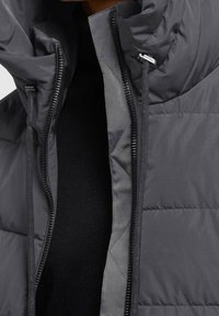 Veste matelassée grise avec un col montant, une fermeture éclair intégrale et un tissu intérieur texturé. Elle présente une surface extérieure lisse et un minimum de mobilier métallique.