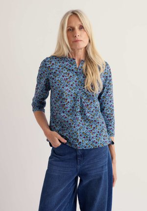 Donna con lunghi capelli biondi indossa una blusa blu a fiori e jeans a gamba larga, in piedi con una mano in tasca su uno sfondo semplice.