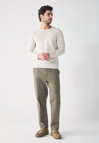 Chemise longue à manches longues beige clair, pantalon vert olive et chaussures marron clair. Design simple avec une coupe décontractée et peu de détails.