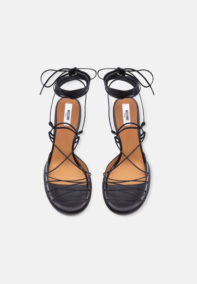 Μαύρα επίπεδα σανδάλια με λεπτά λουράκια που διασταυρώνονται και δεσίματα στον αστράγαλο, ταμπά εσωτερικές σόλες και ετικέτα με το λογότυπο στο εσωτερικό.